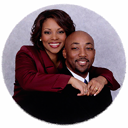 James and Michelle Crowley pose together against a light purple background. Michelle stands behind James with her hands on his shoulders. Both are smiling warmly at the camera, conveying joy and togetherness in a professional portrait setting.