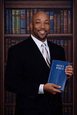 Pastor wearing a dark suit and striped tie standing before a wood-shelved library, holding a blue hardcover book labeled HOLY BIBLE with both hands at chest level; formal, dignified, welcoming tone.
