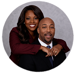 James and Michelle Kelly pose together against a light purple background. Michelle stands behind James with her hands on his shoulders. Both are smiling warmly at the camera, conveying joy and togetherness in a professional portrait setting.