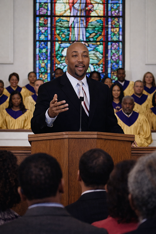 Male pastor standing at a wooden pulpit speaking into a microphone with one hand raised; a church choir in purple and gold robes stands behind him in front of a tall stained glass window depicting Jesus; seated worshippers visible in the foreground facing the pulpit; interior church setting with a reverent, engaged, uplifting tone.